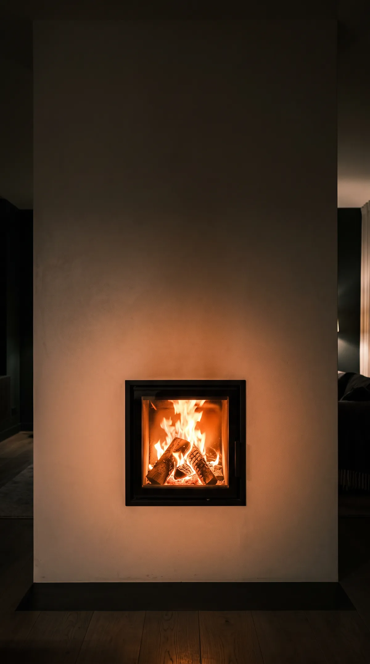 Poêle de masse contemporain, un bloc massif enduit à la chaux dans un séjour sombre du soir, sa petite porte vitrée montrant un feu vif qui éclaire l’enduit